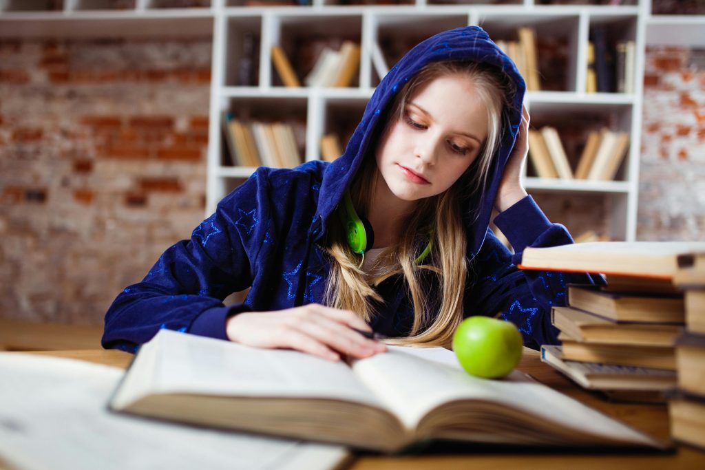 pexels-photo-1326947-1326947 Teenage girl in blue hood reads in library, headphones and apple by her side.