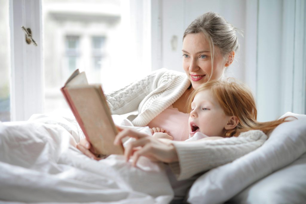 pexels-photo-3755514-3755514 Cheerful young woman hugging cute little girl and reading book together while lying in soft bed in light bedroom at home in daytime